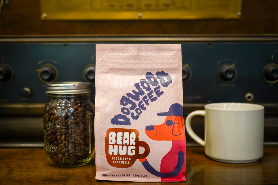 The package of Dogwood Coffee with a cartoon dog on the package, standing by a mason jar of coffee beans and a mug. 